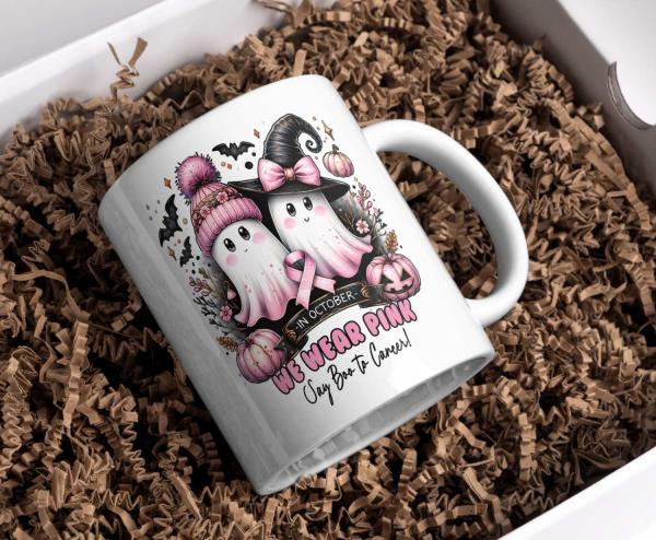 Weiße Halloween-Tasse mit zwei niedlichen Geistern, von denen einer eine rosa Mütze und der andere einen Hexenhut trägt, beide mit rosa Schleife als Symbol für den Kampf gegen Brustkrebs. Mit Kürbissen, Fledermäusen und dem Schriftzug ‚We Wear Pink – Say