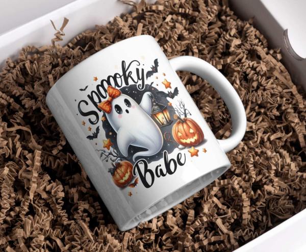 Weiße Tasse mit Halloween-Motiv eines niedlichen Geistes mit orangefarbener Schleife und dem Text ‚Spooky Babe‘. Umgeben von Kürbissen, einer Laterne und Fledermäusen – perfektes Design für Halloween-Liebhaber mit einem Hauch von Niedlichkeit.