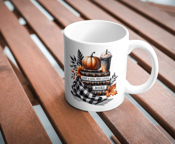 Keramiktasse mit herbstlichem Design und der Aufschrift 'Just a Girl Who Loves Books'. Die Illustration zeigt einen Kürbis, einen Stapel Bücher, ein herbstliches Getränk, einen karierten Schal und bunte Herbstblätter. Die Tasse steht auf einem Holztisch u