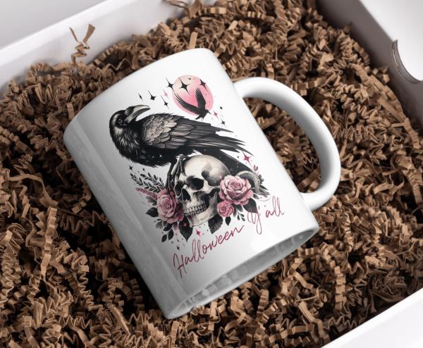 Weiße Tasse mit Halloween-Design: Ein schwarzer Rabe sitzt auf einem Totenkopf, umgeben von rosafarbenen Rosen und Blättern. Im Hintergrund fliegt ein weiterer Rabe vor einem rosa Mond. Der Schriftzug 'Halloween y'all' in elegantem Schriftstil rundet das
