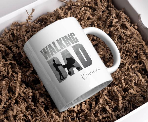 Eine Tasse mit dem personalisierten Schriftzug "The Walking Dad" zeigt eine stilisierte Silhouette eines Vaters mit einem Kinderwagen. Der Name "Kevin" ist ebenfalls auf der Tasse zu sehen. Die Tasse ist auf einem braunen Füllmaterial platziert, was ihr e