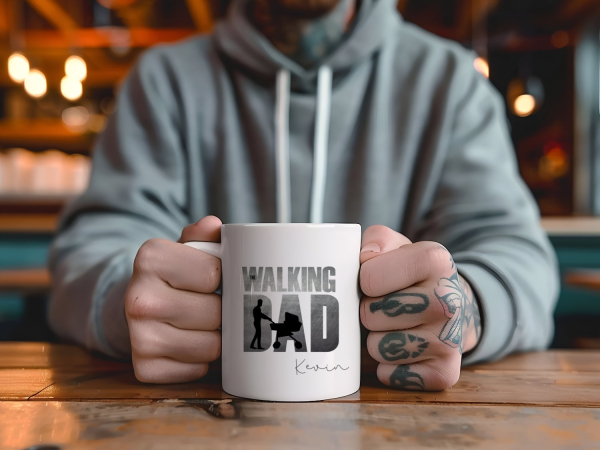 Mann mit Tätowierungen hält eine weiße Tasse mit der Aufschrift „The Walking Dad“ und dem personalisierten Namen „Kevin“, fotografiert in einem gemütlichen Café-Setting.