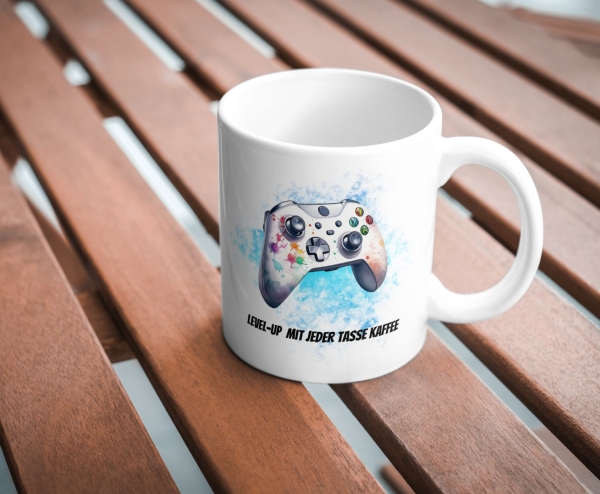 Tasse mit farbigem Gaming-Controller-Design auf blauem Hintergrund, platziert auf einem Holztisch. Der Text unterhalb des Controllers lautet: 'Level-Up mit jeder Tasse Kaffee'. Ein ideales Geschenk für Gamer, die ihre Leidenschaft für Videospiele mit eine