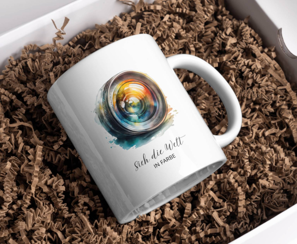 Eine weiße Keramiktasse mit einem farbenfrohen Motiv eines Kameraobjektivs. Das Objektiv ist in lebendigen Farben dargestellt und vermittelt eine künstlerische Atmosphäre. Darunter steht der Schriftzug „sich die Welt in Farbe“ in eleganter Schrift. Diese