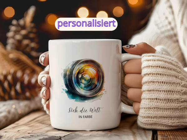 Personalisierte Fotografen-Tasse mit bunter Kameralinse und Schriftzug, gehalten von einer Frau in einem warmen Strickpullover, im gemütlich beleuchteten Winterambiente.