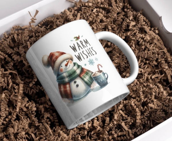 Weißer Keramikbecher mit dem weihnachtlichen Motiv „Warm Wishes“, das einen lächelnden Schneemann mit buntem Schal und Mütze zeigt, neben einer Tasse heißer Schokolade mit Sahne und Zuckerstange, in einer Schachtel mit Holzwolle platziert, bereit als fest