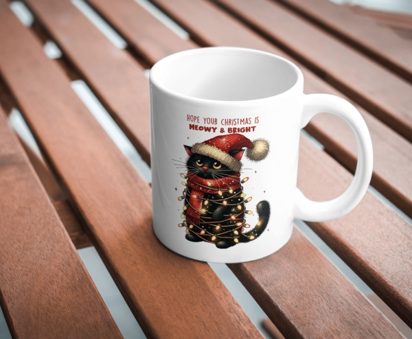 Weiße Tasse mit festlichem Design auf einem Holztisch: Eine schwarze Katze mit roter Weihnachtsmütze und rotem Schal ist in eine Lichterkette gewickelt. Der Text über der Katze lautet 'Hope your Christmas is Meowy & Bright'. Ideales Weihnachtsgeschenk für
