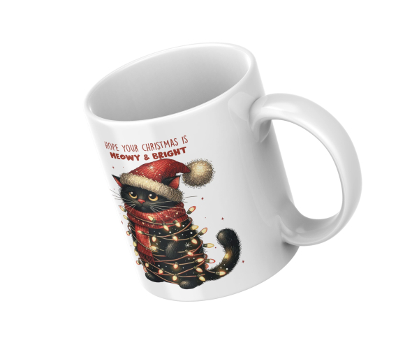 Schräg gestellte weiße Tasse mit festlichem Katzendesign: Eine schwarze Katze mit roter Weihnachtsmütze und rotem Schal, umwickelt von einer Lichterkette. Über der Katze steht der Text 'Hope your Christmas is Meowy & Bright'. Perfekt als Geschenk für Katz