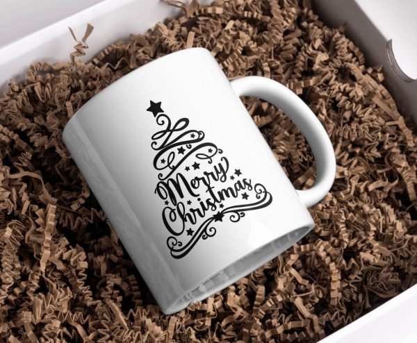 Weiße Keramiktasse mit schwarzem Design eines Weihnachtsbaums in Form der eleganten Aufschrift 'Merry Christmas' im kalligrafischen Stil, platziert in einer Geschenkbox mit Papierfüllung.