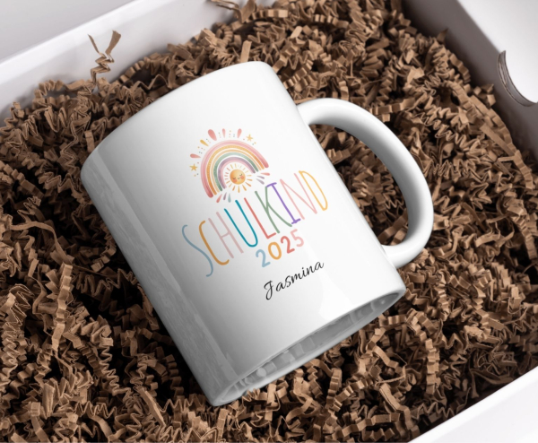 Personalisierte Tasse „Schulkind 2025“ mit buntem Regenbogen und Sonnengesicht, Name „Jasmina“, in Geschenkbox mit brauner Holzwolle arrangiert.