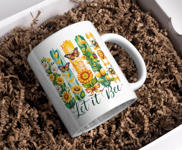 Weiße Keramiktasse mit farbenfrohem Motiv aus Sonnenblumen, Bienen, Schmetterlingen und dem Schriftzug „Let it Bee“, stilvoll präsentiert in Geschenkbox mit brauner Holzwolle.