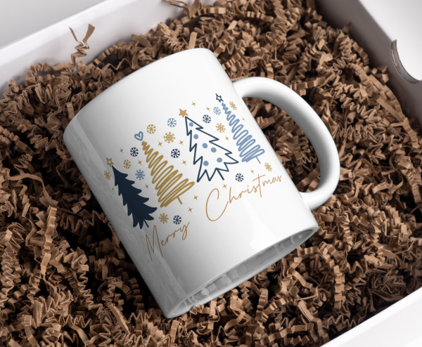 Weihnachtstasse Merry Christmas mit blau-goldenen Weihnachtsbäumen in Geschenkbox auf Papierfüllmaterial.