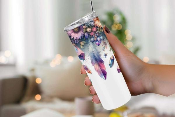 Hand hält einen eleganten Tumbler mit Blumen-, Feder- und Kristalldesign in leuchtenden Farben auf weißem Hintergrund. Hintergrund unscharf, im gemütlichen Wohnzimmerstil.