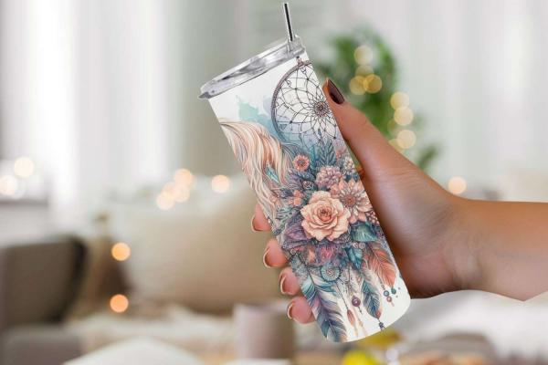 Hand hält einen Reisebecher mit einem detailreichen Design aus Blumen, Federn und Traumfängern. Das Design zeigt zarte Rosatöne, Lila und Pastellfarben mit einem Traumfänger und floralen Elementen, die das Bild dominieren. Der Becher hat einen durchsichti