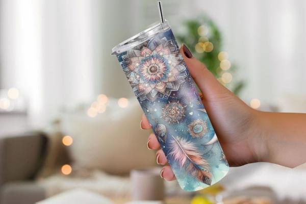 Eine Hand hält einen Thermobecher mit spirituellem Design aus Mandalas, Federn und geometrischen Symbolen in sanften Blau- und Beigetönen. Der Hintergrund ist weich fokussiert und vermittelt eine gemütliche und friedliche Atmosphäre, die das mystische und