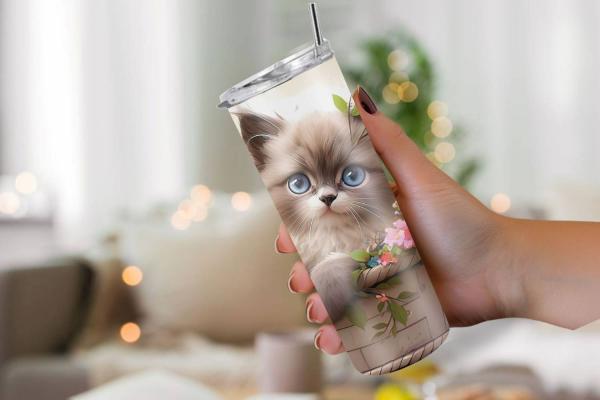 Eine Hand hält einen Thermobecher mit einem niedlichen Design, das ein flauschiges Kätzchen mit großen blauen Augen in einem Korb zeigt, umgeben von bunten Blumen und grünen Blättern. Der Hintergrund ist weich fokussiert und vermittelt eine gemütliche, wo