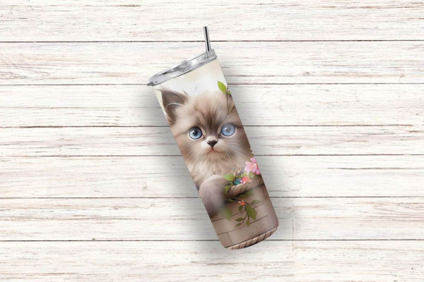 Ein schräg liegender Thermobecher mit einem niedlichen Design, das ein flauschiges Kätzchen mit großen blauen Augen in einem Korb zeigt, dekoriert mit Blumen und grünen Blättern. Das sanfte Design vermittelt eine fröhliche und naturverbundene Atmosphäre –