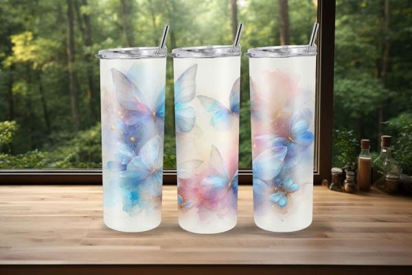 Drei elegante Thermobecher mit einem Design aus pastellfarbenen Schmetterlingen stehen vor einem Fenster, das einen grünen Wald im Hintergrund zeigt. Die Becher sind verziert mit zarten blauen und rosafarbenen Schmetterlingen, die eine luftige und verträu