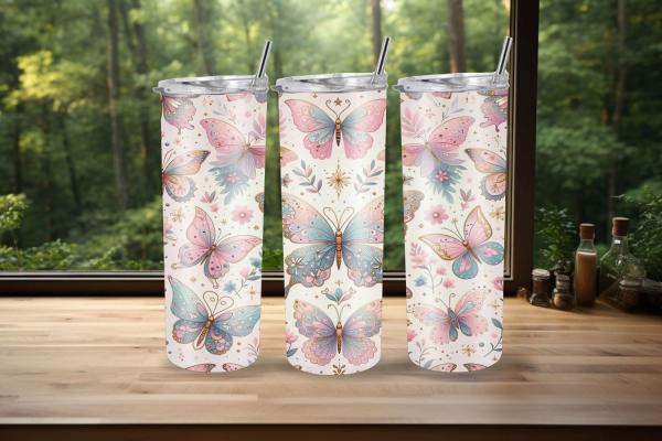 Drei Thermobecher mit pastellfarbenem Schmetterlingsdesign stehen auf einer Holzoberfläche vor einem Fenster mit grünem Waldhintergrund. Das Design zeigt rosa und blaue Schmetterlinge mit zarten Verzierungen und floralen Elementen, die eine fröhliche und