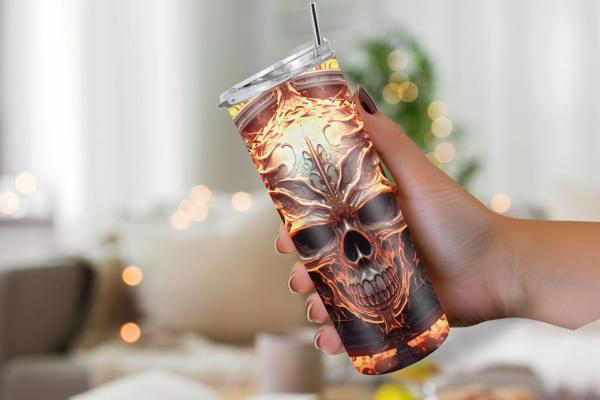 Hand hält einen Thermobecher mit einem metallisch-flammenden Totenkopf-Design in warmen Gelb- und Orangetönen. Der Becher hat einen silbernen Deckel und Strohhalm. Im Hintergrund ist eine unscharfe, gemütliche Wohnzimmerszene mit sanften Lichtern.