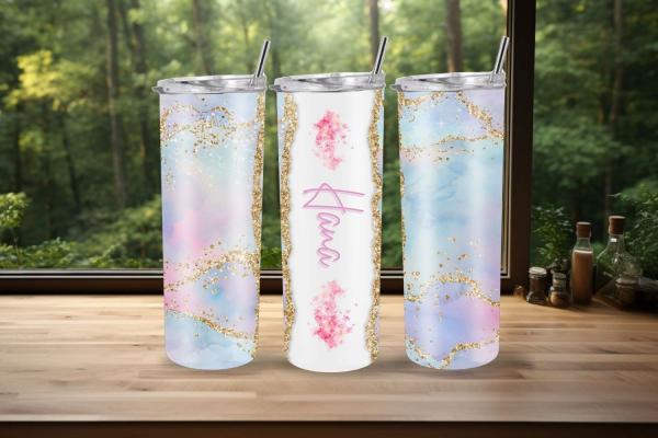Drei elegante Thermobecher auf einer Holzoberfläche vor einem Fenster mit Waldblick. Die äußeren Becher haben ein pastellfarbenes Design in Rosa, Blau und Lila mit goldenen Glitzerakzenten. Der mittlere Becher ist weiß mit dem Namen 'Hana' in pinker Schri