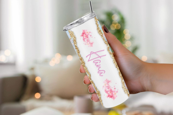 Hand hält einen eleganten weißen Thermobecher mit dem Namen 'Hana' in pinker Schrift. Der Becher ist verziert mit rosa Blumenmotiven und goldenen Glitzerakzenten an den Rändern. Im Hintergrund ist eine unscharfe, gemütliche Wohnzimmerszene mit warmen Boke