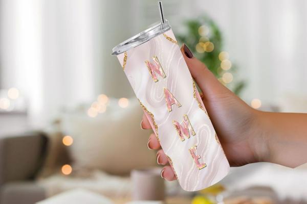 Hand hält einen eleganten Thermobecher mit personalisiertem Design. Der Becher hat einen cremefarbenen Hintergrund mit goldenen Glitzerakzenten und den Schriftzug 'NAME' in großen, roségoldenen Buchstaben. Im Hintergrund ist eine unscharfe, gemütliche Woh