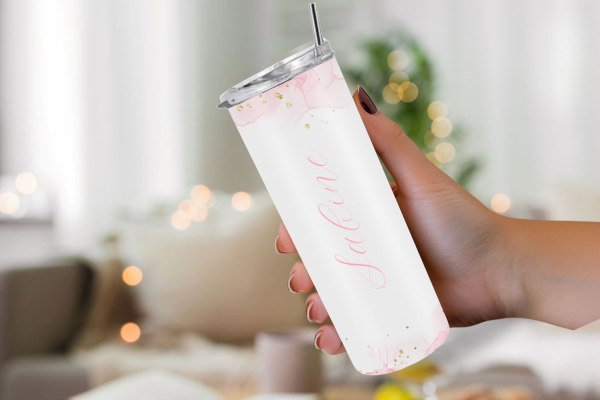 Eine Hand hält einen personalisierten Thermobecher mit dem Namen „Sabine“ in einem zarten rosa Aquarell-Design mit goldenen Akzenten. Der Hintergrund ist weich fokussiert und vermittelt eine gemütliche, einladende Atmosphäre.