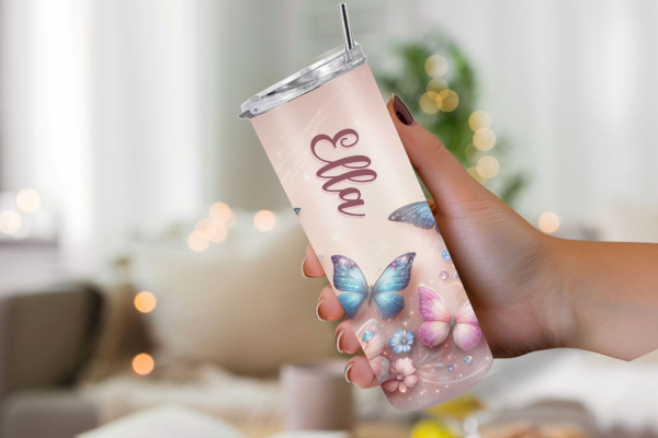 Eine Hand hält einen personalisierten Thermobecher mit dem Namen „Ella“, gestaltet mit einem märchenhaften Schmetterlings- und Blumenmotiv auf einem pastellfarbenen Hintergrund. Der Hintergrund ist weich fokussiert und erzeugt eine gemütliche, traumhafte