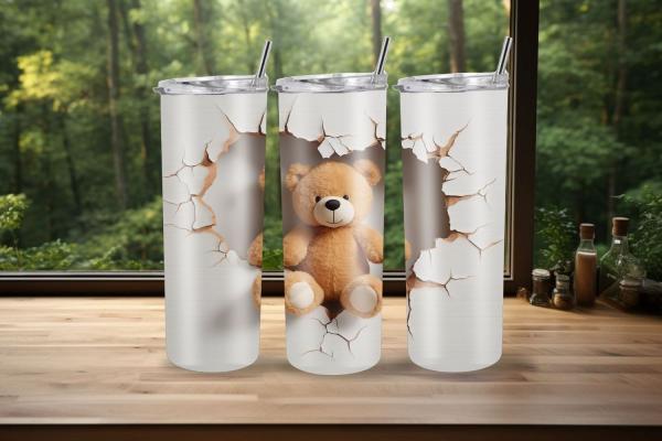 Drei Thermobecher mit einem verspielten Design, das eine zerbrochene Wand zeigt, durch die ein kuscheliger, brauner Teddybär in der Mitte hervorschaut. Die Becher stehen auf einer Holzoberfläche vor einem Fenster mit Blick ins Grüne und haben silberne Dec