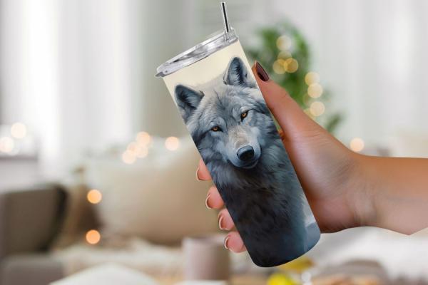 Hand hält einen Thermobecher mit einem detaillierten Wolfsportrait, das das Gesicht eines grauen Wolfs mit durchdringendem Blick zeigt. Der Becher hat einen silbernen Deckel mit Edelstahlstrohhalm. Im Hintergrund ist eine unscharfe, gemütliche Wohnzimmers