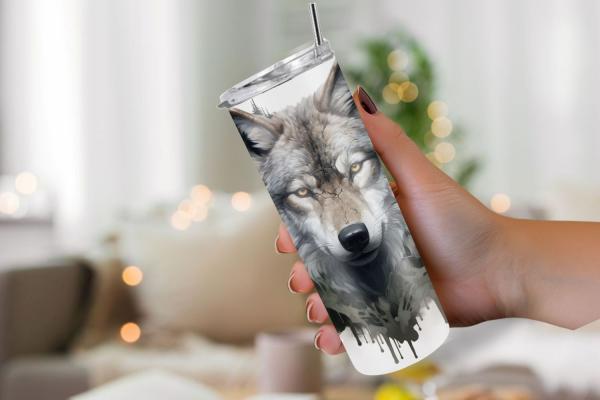 Hand hält einen Thermobecher mit künstlerischem Wolfsmotiv, das das Gesicht eines grauen Wolfs mit intensivem Blick zeigt. Das Design enthält verlaufende Tintenakzente, die einen künstlerischen Wasserfarben-Effekt erzeugen. Der Becher hat einen silbernen