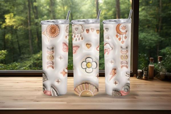 Drei stilvolle Boho-Tumbler mit Sonnen-, Mond- und Blumenmotiven sowie Schmetterling und Herzen, platziert auf einer Holzoberfläche vor einem Fenster mit Waldhintergrund. Perfekt für warme und kalte Getränke.