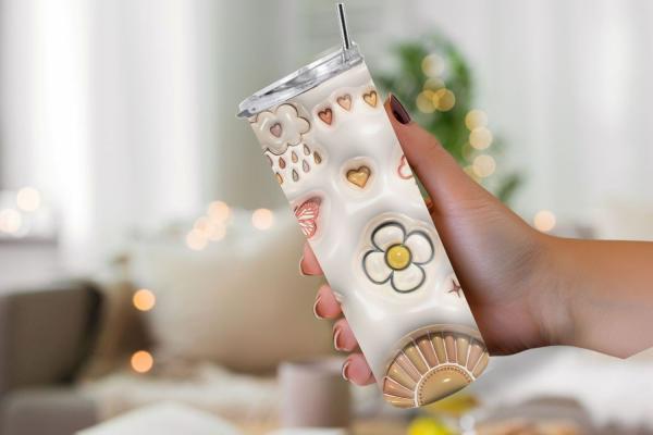 Hand hält einen stilvollen Boho-Tumbler mit Sonnen-, Mond- und Blumenmotiven sowie Herzen und Schmetterling, ideal für warme und kalte Getränke. Hintergrund unscharf, im gemütlichen Wohnzimmerstil.
