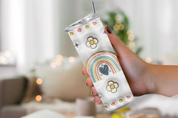 Hand hält einen Boho-Tumbler mit bunten Regenbogen-, Blumen- und Herzmotiven, ideal für warme und kalte Getränke. Hintergrund unscharf, im gemütlichen Wohnzimmerstil.
