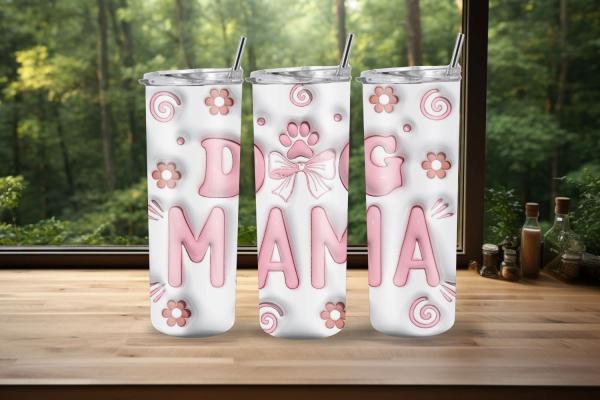 Drei weiße Thermobecher mit der Aufschrift 'DOG MAMA' in rosa Buchstaben, präsentiert vor einem Fenster mit Waldhintergrund. Die Becher sind mit einem Pfotenabdruck, rosa Schleifen, Blumen und Spiralmustern dekoriert. Ideal für Hundeliebhaberinnen.