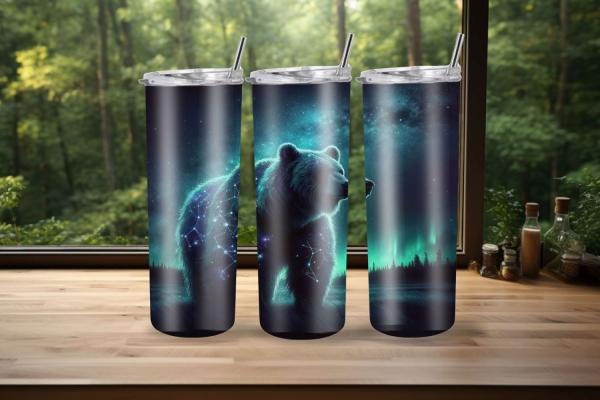 Drei schlanke Reisebecher mit einem majestätischen Bärenmotiv in Blau- und Grüntönen, das Sternbilder und Polarlichter zeigt. Die Becher stehen auf einer Holzoberfläche mit einem Blick auf einen Wald im Hintergrund. Jeder Becher hat einen durchsichtigen D