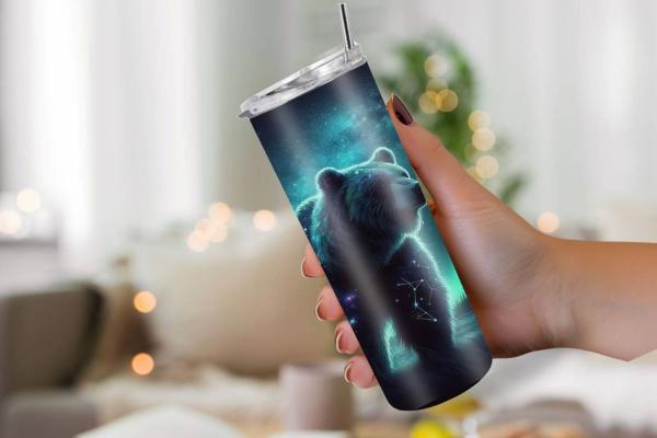 Hand hält einen schlanken Reisebecher mit einem Bärenmotiv, das in leuchtendem Blau und Grün dargestellt ist. Der Bär zeigt Sternbilder und den Nachthimmel mit Polarlichtern. Der Becher hat einen durchsichtigen Deckel und einen Edelstahlstrohhalm, ideal f