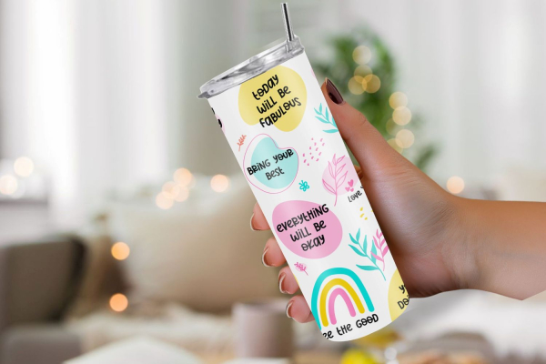 Hand hält einen weißen Thermobecher mit farbenfrohen, motivierenden Sprüchen wie 'Today will be fabulous', 'Bring your best' und 'Everything will be okay'. Der Becher ist mit Illustrationen von Regenbogen und Blättern dekoriert. Im Hintergrund ist ein gem