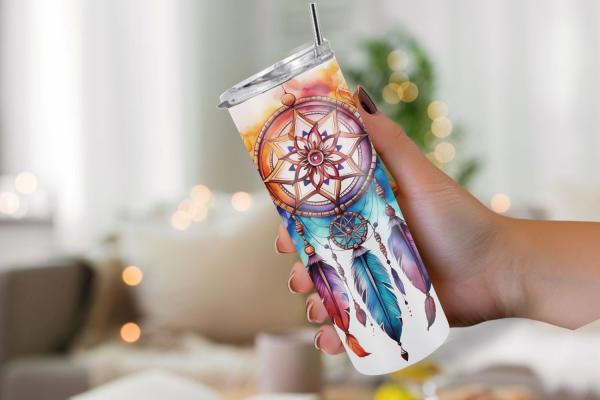 Hand hält einen Thermobecher mit detailreichem Traumfänger-Design, verziert mit bunten Federn und floralen Elementen im Boho-Stil. Das Bild vermittelt eine gemütliche Atmosphäre, perfekt für Liebhaber von Boho-Accessoires.