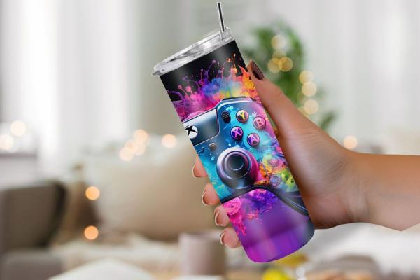 Hand hält einen Thermobecher mit lebendigem Gaming-Controller-Design auf schwarzem Hintergrund, umgeben von spritzenden, bunten Farbakzenten. Ein auffälliges und dynamisches Design, perfekt für Gaming-Fans.