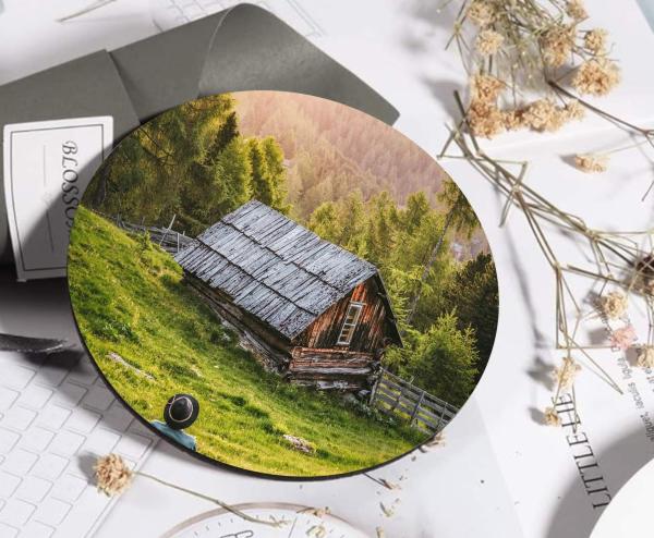 Runder Untersetzer mit einem Bild einer alten, rustikalen Holzhütte in einer grünen, hügeligen Landschaft. Im Vordergrund ist eine Person mit Hut von hinten zu sehen, die zur Hütte und dem umgebenden Wald blickt. Der Untersetzer liegt auf einem Schreibtis