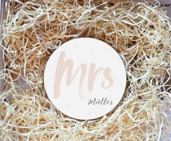 Runder Untersetzer mit dem Schriftzug „Mrs“ in beigefarbener Schrift und dem Nachnamen „Müller“ darunter in eleganter Schreibschrift. Der Untersetzer liegt in einer Geschenkbox, umgeben von natürlichem Holzwolle-Füllmaterial. Perfekt als personalisiertes