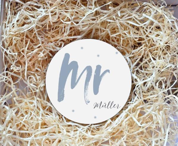 Runder Untersetzer mit dem Schriftzug „Mr“ in graublauer Schrift und dem Nachnamen „Müller“ darunter in eleganter Schreibschrift. Der Untersetzer liegt in einer Geschenkbox, umgeben von natürlichem Holzwolle-Füllmaterial. Perfekt als personalisiertes Gesc
