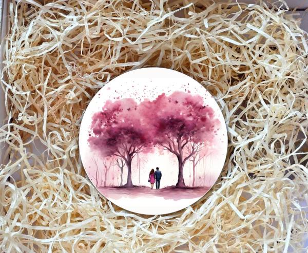 Runder Untersetzer mit romantischer Illustration von zwei Personen, die unter rosa blühenden Bäumen spazieren gehen. Der Untersetzer liegt in einer Geschenkbox, umgeben von natürlichem Holzwolle-Füllmaterial. Ideal als personalisiertes Geschenk für Paare