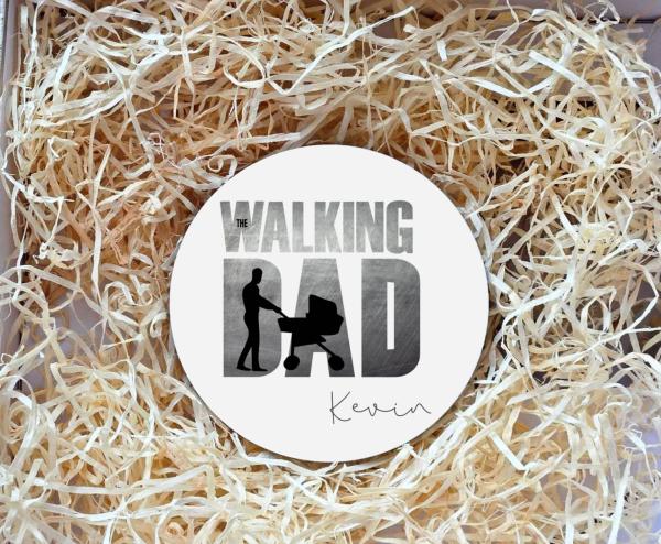 Runder Untersetzer mit der humorvollen Aufschrift „The Walking Dad“ und einer Silhouette eines Vaters, der einen Kinderwagen schiebt. Unter dem Motiv steht der Name „Kevin“ in eleganter Schrift. Der Untersetzer liegt in einer Geschenkbox, umgeben von natü